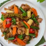 A plate of Pad Kee Mao / Drunken Noodles with rice noodles, chicken, peppers, baby corn, carrots, long beans, young green peppercorns, and holy basil from the top down view