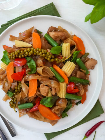 A plate of Pad Kee Mao / Drunken Noodles with rice noodles, chicken, peppers, baby corn, carrots, long beans, young green peppercorns, and holy basil from the top down view